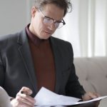 Smart Contracts - Elegant adult man in jacket and glasses looking through documents while sitting on white sofa in luxury room