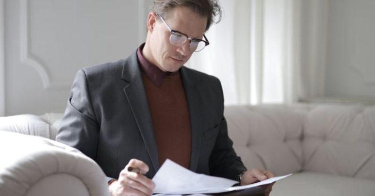 Smart Contracts - Elegant adult man in jacket and glasses looking through documents while sitting on white sofa in luxury room