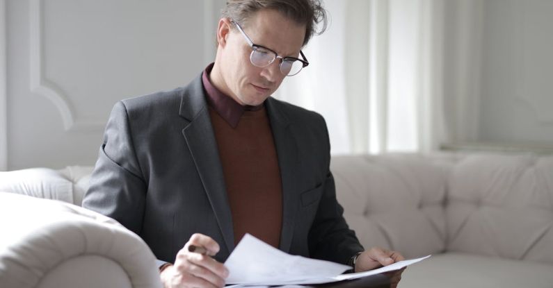 Smart Contracts - Elegant adult man in jacket and glasses looking through documents while sitting on white sofa in luxury room